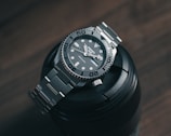 A sleek and polished stainless steel wristwatch with a robust bezel and luminescent hour markers is resting atop a black camera lens on a wooden surface. The watch displays the brand name and has a neatly linked metallic strap.
