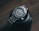 A sleek and polished stainless steel wristwatch with a robust bezel and luminescent hour markers is resting atop a black camera lens on a wooden surface. The watch displays the brand name and has a neatly linked metallic strap.