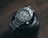 A sleek and polished stainless steel wristwatch with a robust bezel and luminescent hour markers is resting atop a black camera lens on a wooden surface. The watch displays the brand name and has a neatly linked metallic strap.
