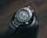 A sleek and polished stainless steel wristwatch with a robust bezel and luminescent hour markers is resting atop a black camera lens on a wooden surface. The watch displays the brand name and has a neatly linked metallic strap.