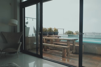 A modern indoor space with a wicker chair and a floor lamp is visible inside. Large glass doors open to an outdoor area featuring a wooden picnic table surrounded by potted plants. A swimming pool is seen in the background, creating a peaceful and inviting atmosphere.