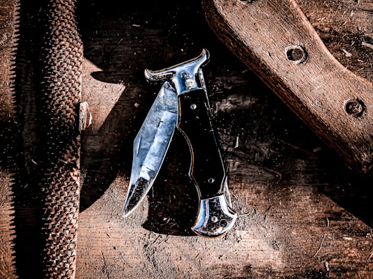 A folding knife with a reflective metallic blade and a black handle lies on a worn wooden surface. To the left of the knife, there is a textured metal file, and on the right side, there is a piece of wood with two circular holes. The surface appears stained and used, giving an old and rustic atmosphere.