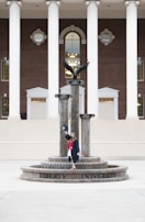 A person in a graduation gown and cap stands on a decorative fountain in front of a large building with tall white columns and a brick facade. A statue of an eagle with its wings spread is positioned atop the fountain.