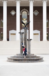 A person in a graduation gown and cap stands on a decorative fountain in front of a large building with tall white columns and a brick facade. A statue of an eagle with its wings spread is positioned atop the fountain.