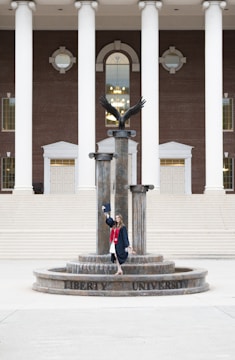 A person in a graduation gown and cap stands on a decorative fountain in front of a large building with tall white columns and a brick facade. A statue of an eagle with its wings spread is positioned atop the fountain.