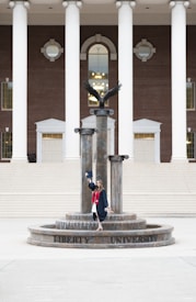 A person in a graduation gown and cap stands on a decorative fountain in front of a large building with tall white columns and a brick facade. A statue of an eagle with its wings spread is positioned atop the fountain.