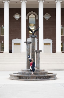 A person in a graduation gown and cap stands on a decorative fountain in front of a large building with tall white columns and a brick facade. A statue of an eagle with its wings spread is positioned atop the fountain.