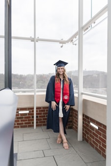 A person is standing in a glass enclosure wearing a graduation cap and gown. The gown is navy blue with a red stole. They are standing on a stone floor with brick and glass walls surrounding them. The sky outside is overcast, with a hint of green landscape in the background.