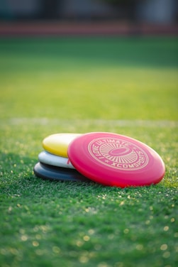A stack of colorful frisbees lies on a lush green grass field. The top frisbee is bright red with a white emblem and text stating 'LOVE ULTIMATE XCOMSPORTS.' Underneath are frisbees in yellow, white, and black hues. The scene is softly lit, suggesting either early morning or late afternoon.