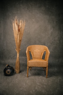 A woven wicker chair is positioned beside a tall arrangement of dried reeds and a dark, round gourd vase against a textured, neutral backdrop, creating a rustic and minimalistic setting.