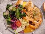 A plate filled with a fresh salad, shrimp topped with garlic and herbs, and rice balls. The salad includes mixed greens, cherry tomatoes, and red cabbage. The shrimp are bright orange and surrounded by a sauce.