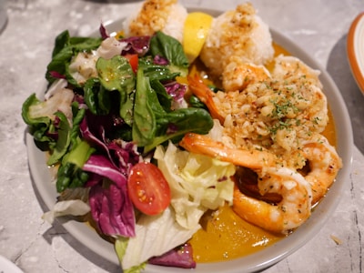 A plate filled with a fresh salad, shrimp topped with garlic and herbs, and rice balls. The salad includes mixed greens, cherry tomatoes, and red cabbage. The shrimp are bright orange and surrounded by a sauce.