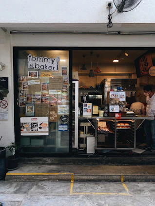 The photo displays a small bakery with a glass door and window showcasing numerous menu items and advertisements. Inside, various baked goods can be seen displayed on the counter. Pendant lights hang from the ceiling, and there is a man wearing a white shirt and mask working behind the counter. Potted plants are located outside the bakery.