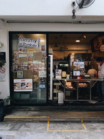 The photo displays a small bakery with a glass door and window showcasing numerous menu items and advertisements. Inside, various baked goods can be seen displayed on the counter. Pendant lights hang from the ceiling, and there is a man wearing a white shirt and mask working behind the counter. Potted plants are located outside the bakery.