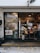 The photo displays a small bakery with a glass door and window showcasing numerous menu items and advertisements. Inside, various baked goods can be seen displayed on the counter. Pendant lights hang from the ceiling, and there is a man wearing a white shirt and mask working behind the counter. Potted plants are located outside the bakery.