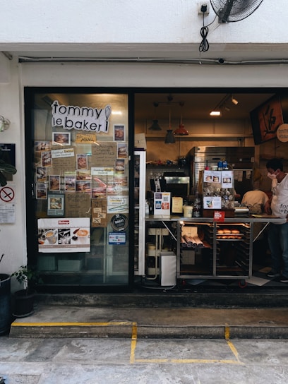 The photo displays a small bakery with a glass door and window showcasing numerous menu items and advertisements. Inside, various baked goods can be seen displayed on the counter. Pendant lights hang from the ceiling, and there is a man wearing a white shirt and mask working behind the counter. Potted plants are located outside the bakery.