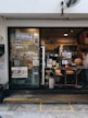 The photo displays a small bakery with a glass door and window showcasing numerous menu items and advertisements. Inside, various baked goods can be seen displayed on the counter. Pendant lights hang from the ceiling, and there is a man wearing a white shirt and mask working behind the counter. Potted plants are located outside the bakery.