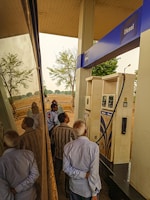 Several people are gathered around a diesel fuel pump at a gas station. The setting includes clear reflections on a shiny surface, likely a vehicle. There are trees and an open field in the background, suggesting a rural location.