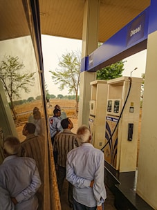 Several people are gathered around a diesel fuel pump at a gas station. The setting includes clear reflections on a shiny surface, likely a vehicle. There are trees and an open field in the background, suggesting a rural location.