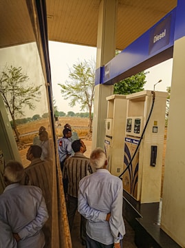 Several people are gathered around a diesel fuel pump at a gas station. The setting includes clear reflections on a shiny surface, likely a vehicle. There are trees and an open field in the background, suggesting a rural location.