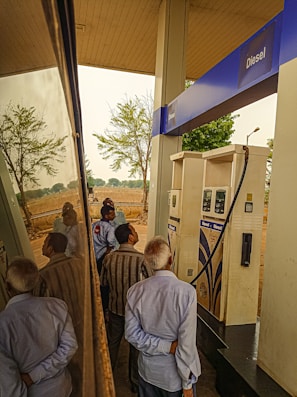 Several people are gathered around a diesel fuel pump at a gas station. The setting includes clear reflections on a shiny surface, likely a vehicle. There are trees and an open field in the background, suggesting a rural location.