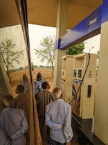 Several people are gathered around a diesel fuel pump at a gas station. The setting includes clear reflections on a shiny surface, likely a vehicle. There are trees and an open field in the background, suggesting a rural location.
