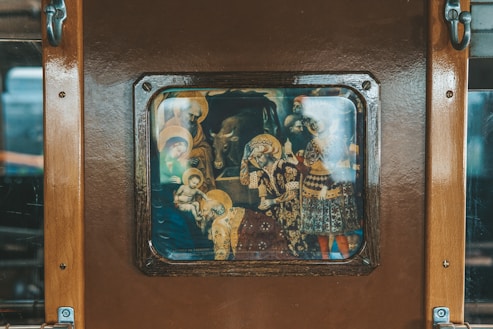 An image depicts a framed artwork mounted on a wooden surface. The artwork is a religious painting featuring multiple figures, including halos, an infant, and a cow. The colors are rich and the figures are dressed in ornate clothing.