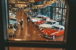 A well-lit showroom displays a collection of classic cars arranged in rows. Two people are examining one of the cars while several others walk around the exhibit. The setting includes reflective glass walls and a suspended vintage airplane, adding to the automotive theme.
