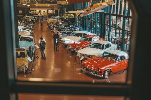A well-lit showroom displays a collection of classic cars arranged in rows. Two people are examining one of the cars while several others walk around the exhibit. The setting includes reflective glass walls and a suspended vintage airplane, adding to the automotive theme.