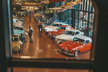 A well-lit showroom displays a collection of classic cars arranged in rows. Two people are examining one of the cars while several others walk around the exhibit. The setting includes reflective glass walls and a suspended vintage airplane, adding to the automotive theme.