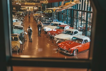 A well-lit showroom displays a collection of classic cars arranged in rows. Two people are examining one of the cars while several others walk around the exhibit. The setting includes reflective glass walls and a suspended vintage airplane, adding to the automotive theme.