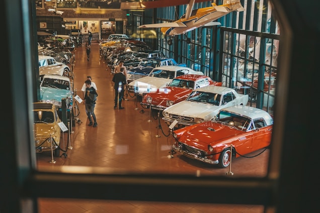 A well-lit showroom displays a collection of classic cars arranged in rows. Two people are examining one of the cars while several others walk around the exhibit. The setting includes reflective glass walls and a suspended vintage airplane, adding to the automotive theme.
