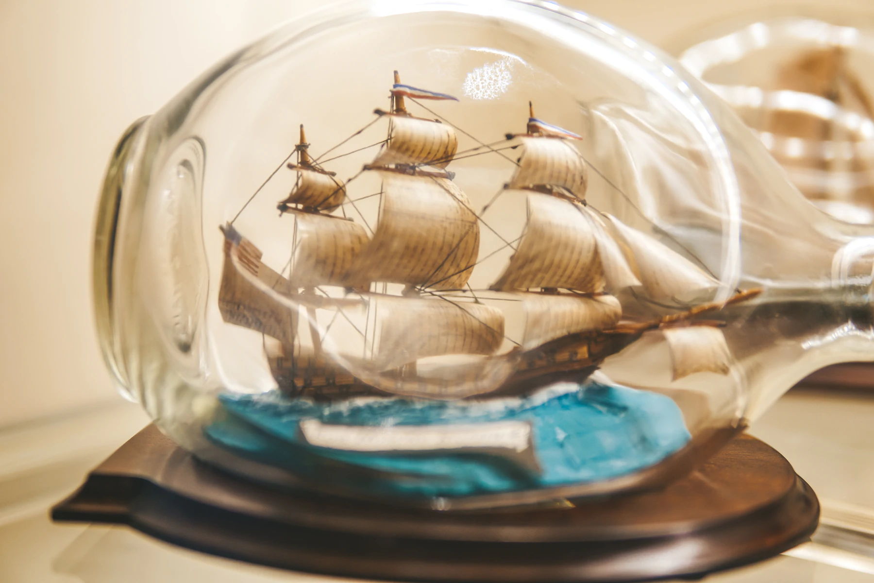 A three-masted model ship under full sail on a blue sea inside a round glass bottle on a dark wooden base