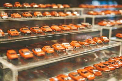A collection of miniature red car models displayed on multiple clear, tiered shelves. The cars are neatly arranged in rows and are all similar in shade, creating a visually cohesive and organized appearance.