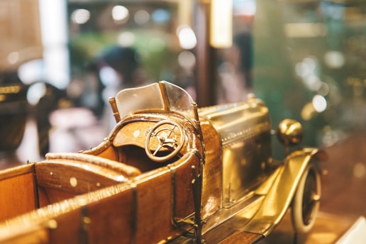A detailed model of a vintage car is prominently displayed, showcasing the intricate craftsmanship. The focus is on the wooden steering wheel and the driver's seat with leather-like textures. The brass finish on the car body reflects ambient light, giving it a polished look.