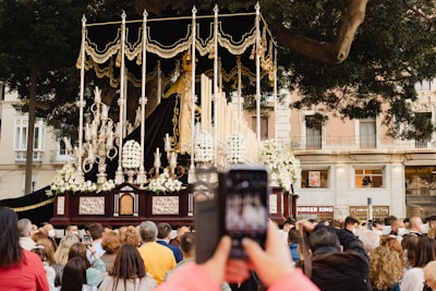 A religious procession with an ornate float featuring a statue surrounded by gold and silver elements, flowers, and tall candles. A crowd of people gathers to watch, with someone in the foreground capturing the scene with a smartphone. The procession takes place in an urban setting with trees and a building in the background.