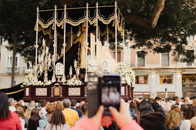 A religious procession with an ornate float featuring a statue surrounded by gold and silver elements, flowers, and tall candles. A crowd of people gathers to watch, with someone in the foreground capturing the scene with a smartphone. The procession takes place in an urban setting with trees and a building in the background.