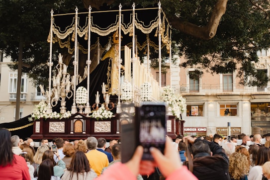 A religious procession with an ornate float featuring a statue surrounded by gold and silver elements, flowers, and tall candles. A crowd of people gathers to watch, with someone in the foreground capturing the scene with a smartphone. The procession takes place in an urban setting with trees and a building in the background.