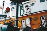 A close-up of a colorful vintage ship featuring white and orange hues with circular windows and a life preserver mounted on the side. The ship is adorned with maritime equipment including gears and a lamp post, suggesting it might be docked. The plaque on the side indicates restoration by a shipyard.