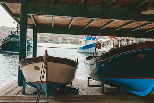 Two wooden boats are docked under a wooden canopy next to a body of water, with more boats visible in the background. A Turkish flag is hanging on one of the boats. The shoreline is in the distance, with buildings and a hilly landscape.
