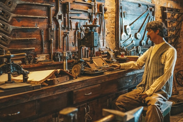 A workshop filled with a variety of vintage tools hanging on the wall. A mannequin dressed in vintage attire is seated, appearing as if working with the machinery on the wooden table. The workshop has a rustic feel with wooden shelves and brick walls.
