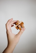 A hand gently holds a small, intricate wooden puzzle against a plain, light background. The puzzle consists of interlocking wooden pieces, suggesting complexity and craftsmanship.