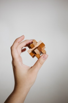 A hand gently holds a small, intricate wooden puzzle against a plain, light background. The puzzle consists of interlocking wooden pieces, suggesting complexity and craftsmanship.