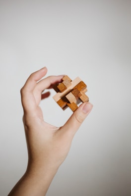 A hand gently holds a small, intricate wooden puzzle against a plain, light background. The puzzle consists of interlocking wooden pieces, suggesting complexity and craftsmanship.
