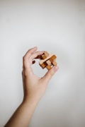 A hand holding a small wooden puzzle against a plain background. The puzzle consists of interlocking wooden pieces, creating a three-dimensional shape. The hand is positioned in a gentle grasp, showcasing the simplicity and craftsmanship of the puzzle.