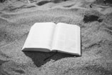 An open book lies on a surface of sand, with visible text and a fabric bookmark hanging out.