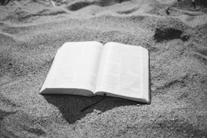 An open book lies on a surface of sand, with visible text and a fabric bookmark hanging out.