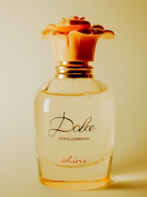 A clear glass perfume bottle with a floral-themed cap that resembles a blossoming flower. The label on the bottle reads 'Dolce & Gabbana' with the word 'shine' in a delicate script near the base. The overall design is elegant with gold accents.
