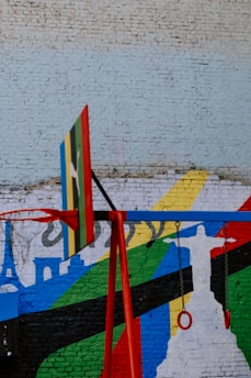 A vibrantly colored mural is painted on a brick wall, depicting a silhouette of the Christ the Redeemer statue against a backdrop of abstract shapes in green, blue, yellow, black, and red stripes. In the foreground, a red swing set with a partial basketball hoop can be seen.