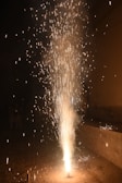 An intense burst of fireworks erupts from the ground, emitting a cascade of bright sparks and trails of light against a dark background. The fiery spectacle illuminates the immediate surroundings, creating a visually captivating display.